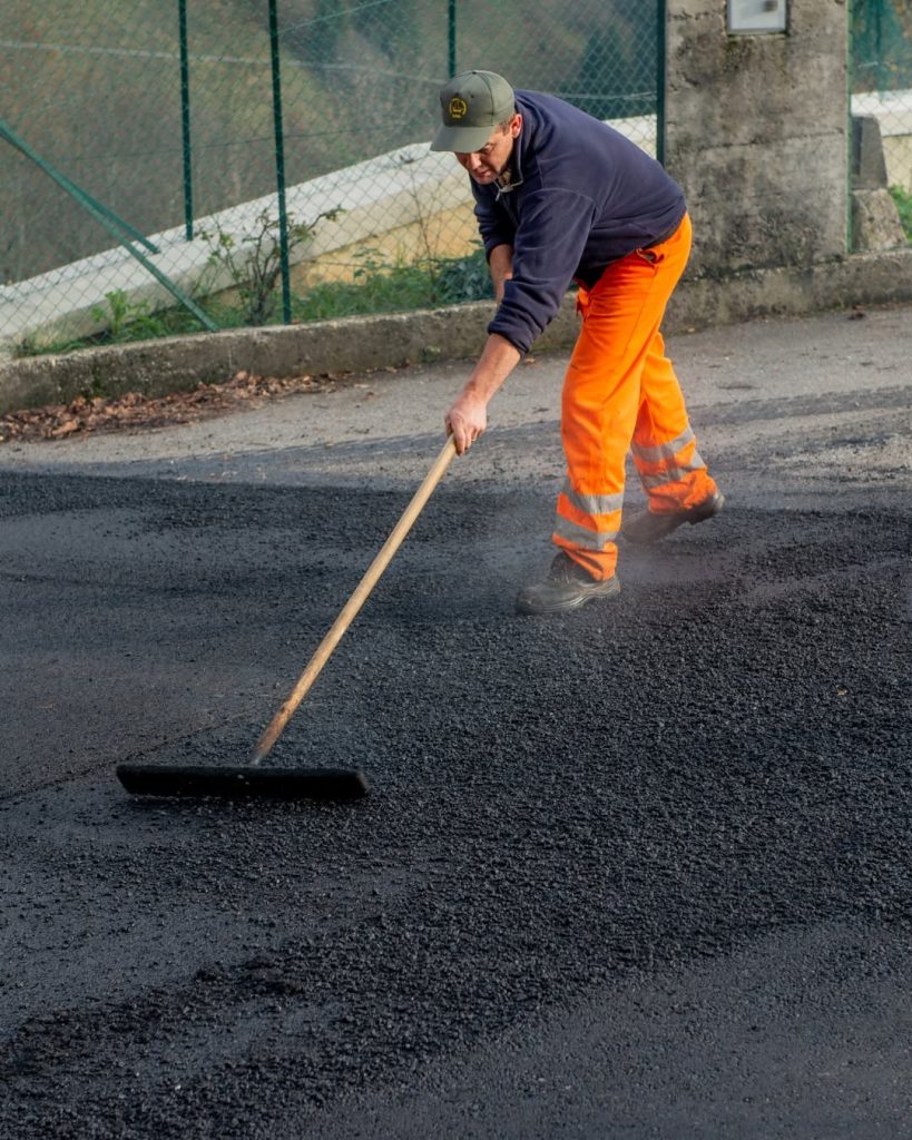 Worker in orange trousers using a long-handled tool to spread and finish fresh asphalt